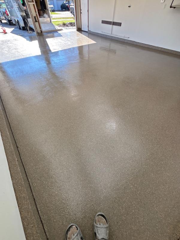 A newly coated, glossy garage floor with an open door, revealing a vehicle and person outside in daylight. Tan-colored surface, sandals visible.